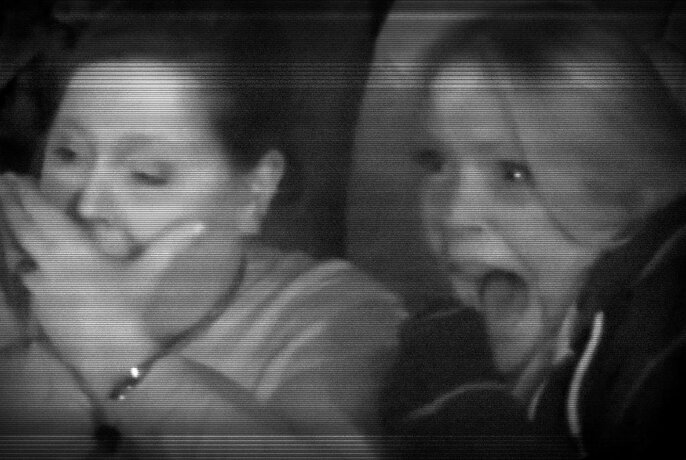 Black and white image of audience members watching something and looking scared, one screaming with mouth open and one covering her mouth with her hand.