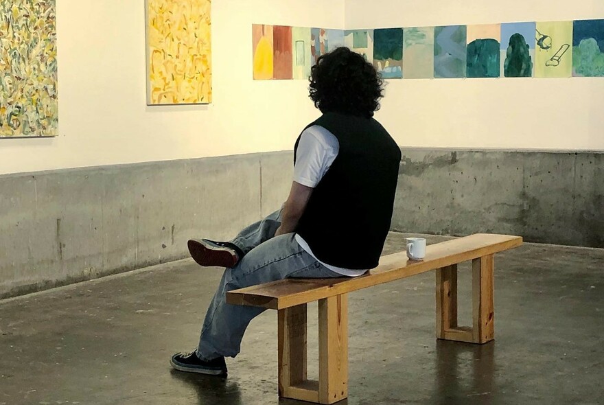 Person sitting on a bench seat inside a gallery looking at the art works on the wall.