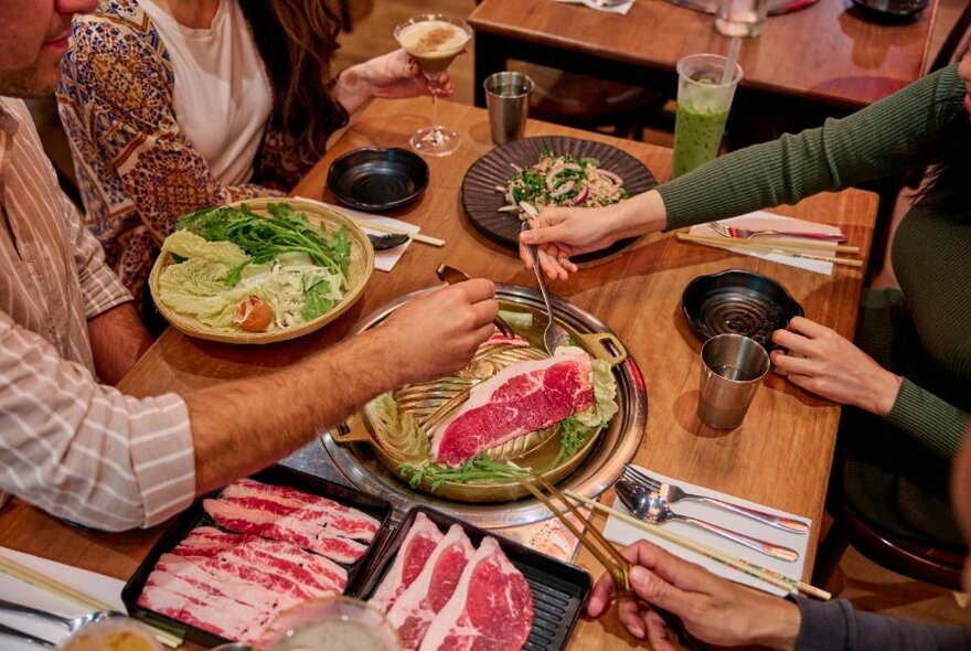 Overhead view of people's hands and arms stretched out and barbequing slices of meat on a hot plate at their restaurant table, with other plates of food, cutlery and drinks nearby on the table.