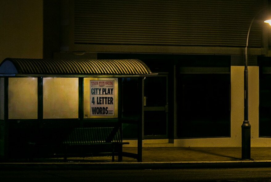 A lonely looking bus shelter at night lit by street lights with a bill poster on the right side. 