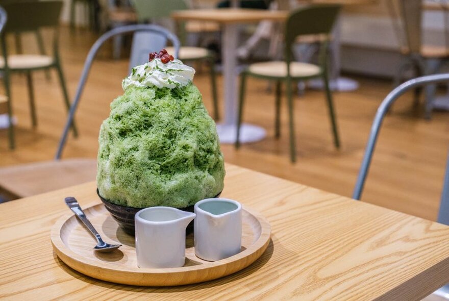 Green shaved ice dessert on a tray with a spoon and cups in a cafe.