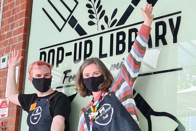 Two people in black masks and aprons, with arms upraised outside Pop-Up Library signed window.