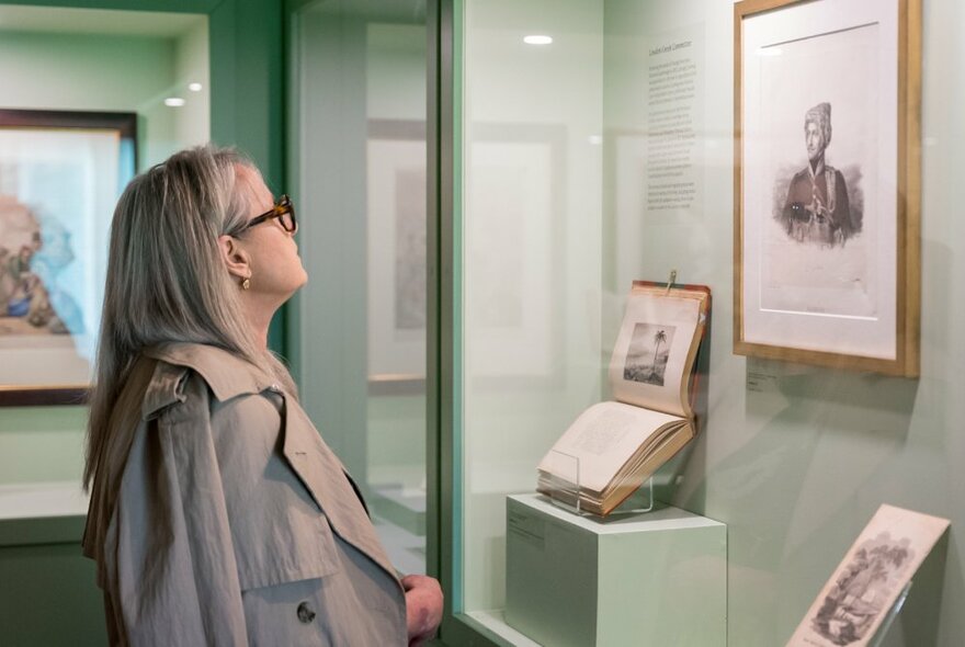 A person looking at a framed portrait, book and image in a display case in an exhibition.
