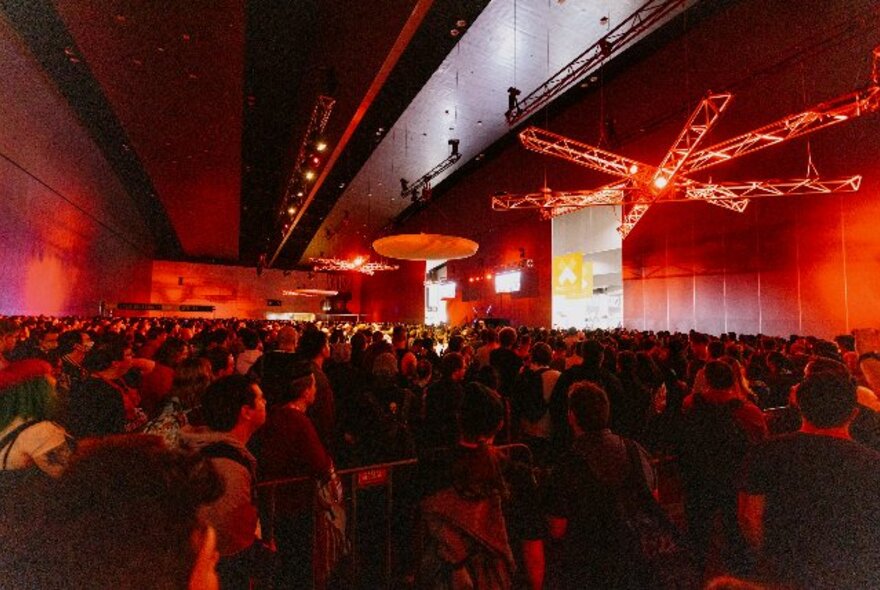 Large crowds in a busy red-lit venue looking towards a screen on a wall.