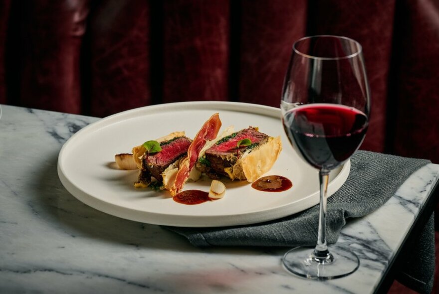 Glass of red wine to right of white plate with elegantly plated meal, on grey marble-look surface.