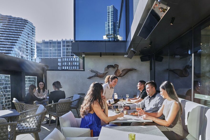 A group of seated people enjoying glasses of wine on the outdoor terrace at Fleet Rooftop Bar, with the Melbourne city skyline in the background.