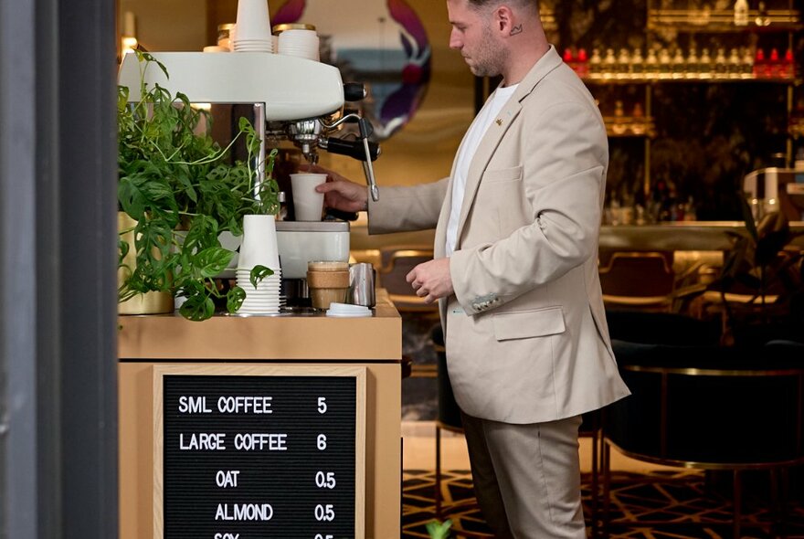 A man wearing a suit making coffee at a small coffee machine located inside the Jin Bar, with a small sign in front advertising the types and prices of coffee for sale.