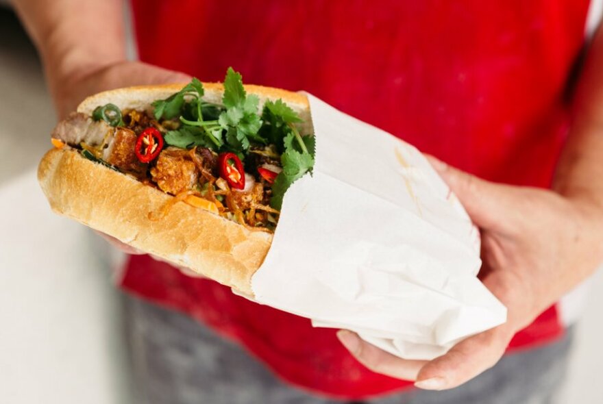 Person holding a Vietnamese banh mi roll with meat, salad and chilli.