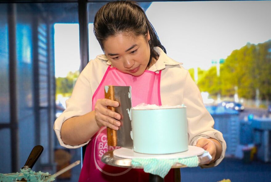 A woman spreading icing on the sides of her pale blue cake with a long spatula, outdoors. 