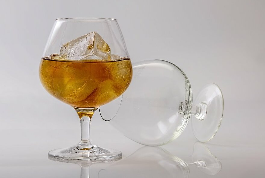 Two brandy balloons, one on its side empty, the other containing spirits and ice.