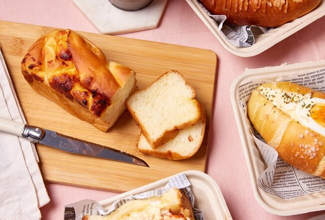 A pink table with a sliced shokupan loaf on a board, surrounded by other bread dishes.