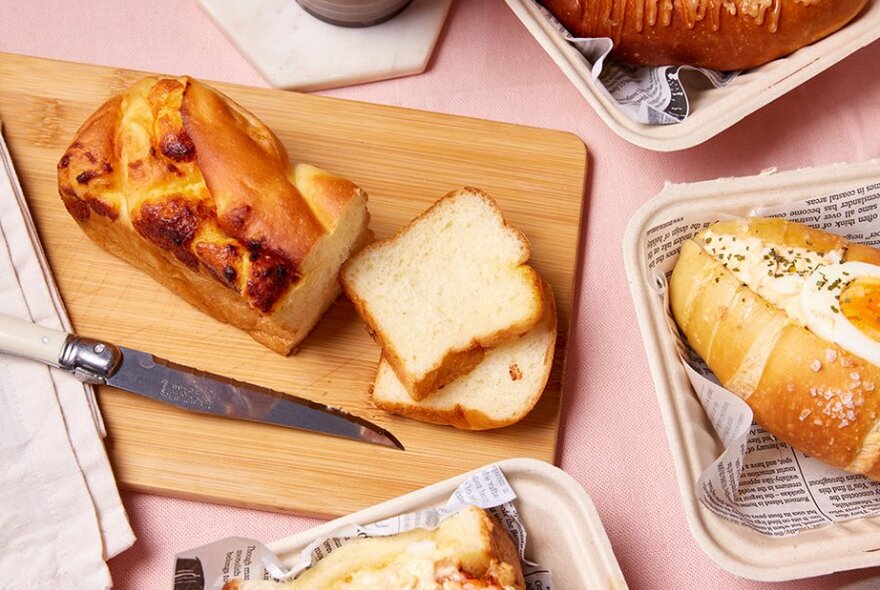 A pink table with a sliced shokupan loaf on a board, surrounded by other bread dishes. 