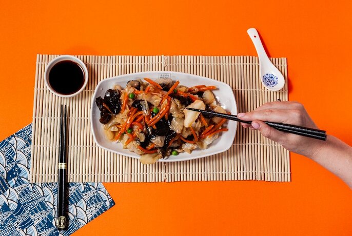 Orange table with a bowl of Korean noodles, bamboo mat, hands holding chopsticks and dipping sauce.