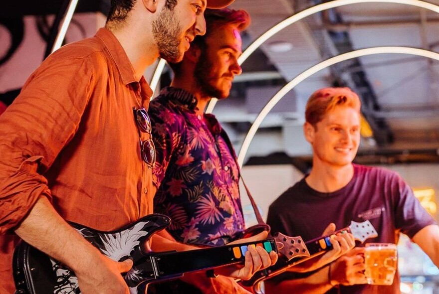 Three mean standing around smiling and drinking beer, two of them holding Guitar Hero game guitars.