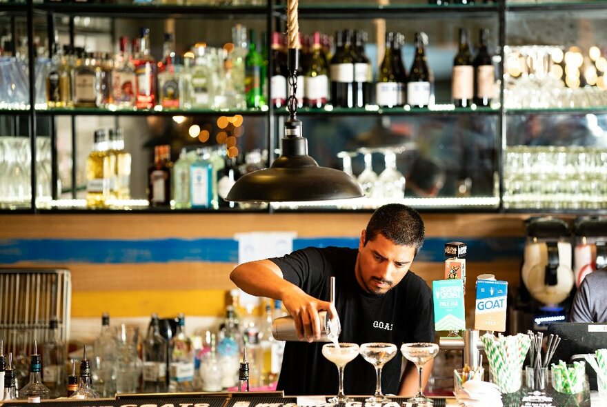 A person behind a bar pouring three margarita drinks from a shaker into cocktail glasses that rest on the bar counter, a shelf of bottles and glasses on the rear wall.