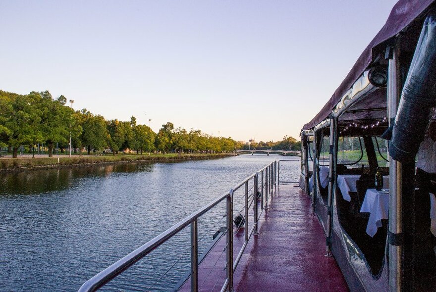 The walkway on the side of a floating river cruise restaurant, with the tree-lined riverbank in the distance.