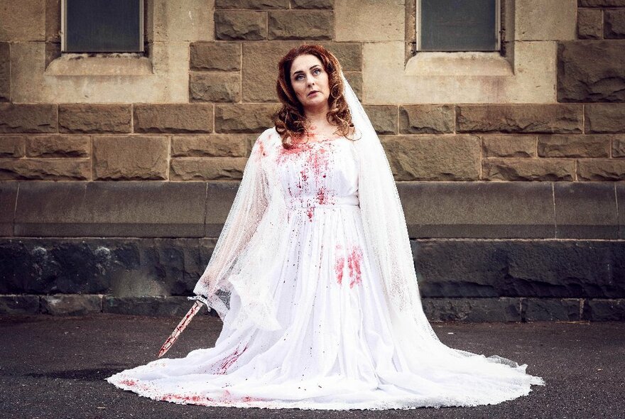 A woman in a white wedding dress and veil, kneeling on the street in front of a bluestone building, with blood on her dress and a bloodied knife in her hand.
