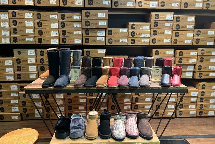 A table display of colourful Ugg boots and Ugg slippers in front of shoeboxes in shelves, in a store setting.