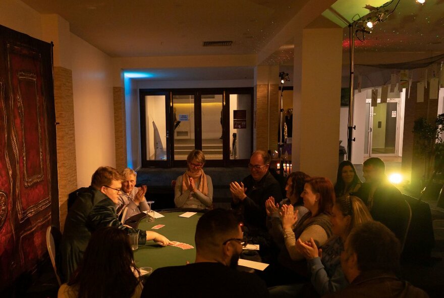 A group of people seated at a green beize table watching a magician perform card tricks.