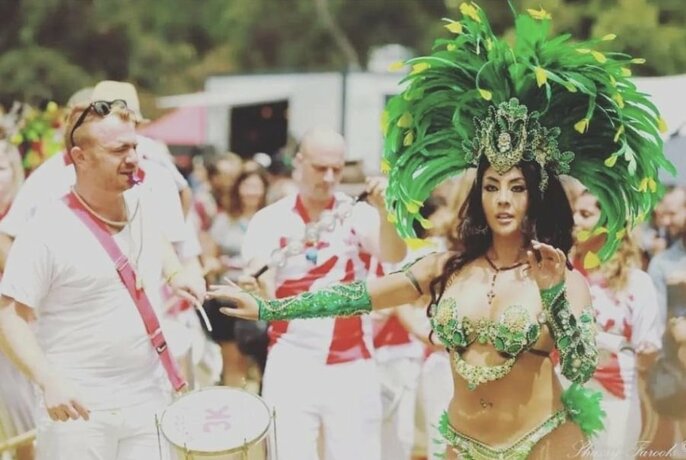 A woman wearing a Latin American dancing costume with a large green feather headdress, dances in front of a line of men in white uniforms, one wearing a drum.