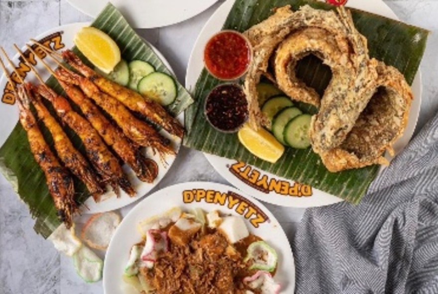 Overhead view of three plates of food on a table including satays, fried fish and a rice dish.