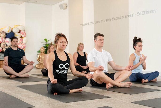 Interior of a yoga studio with six participants sitting cross-legged on mats on the floor.