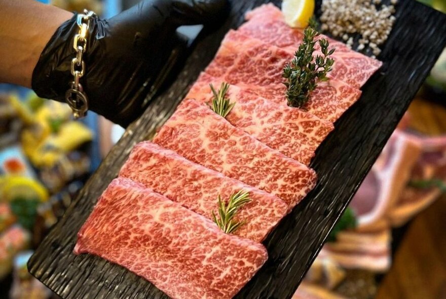 A plate displaying thin slices of wagyu beef being held by a hand wearing a black kitchen glove. 