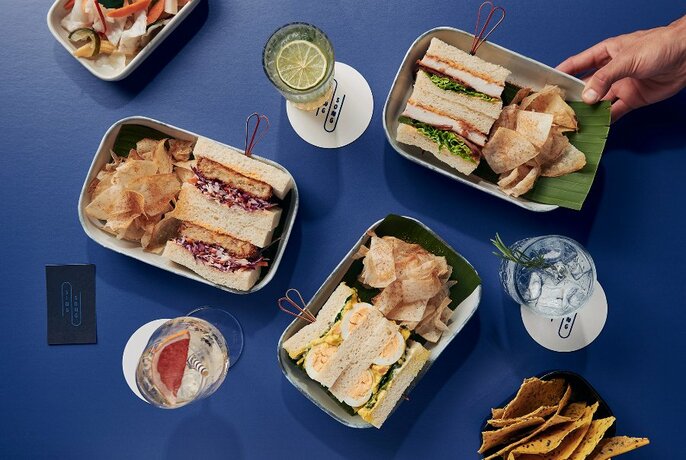 Overhead view of plates of assorted sandos with varied fillings, plus crisps and drinks on coasters.