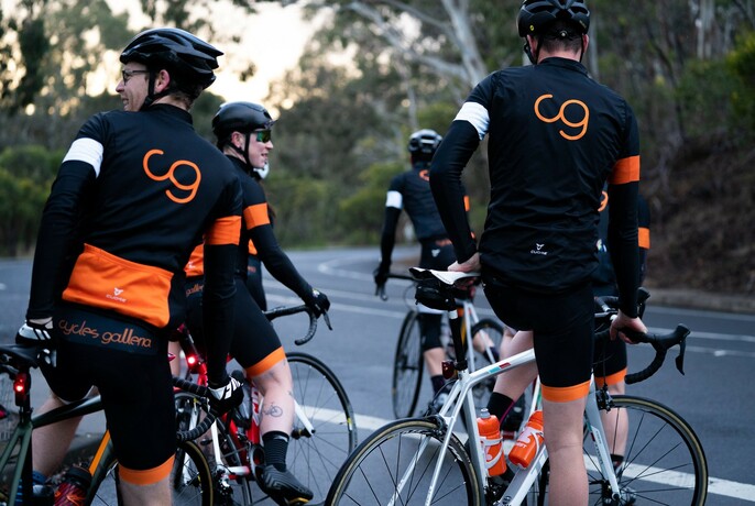 A group of men all wearing branded Cycle Galleria lycra riding gear straddling their bikes as if about to commence riding.