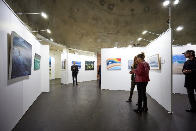 Installation view of the maritime exhibition with a few people viewing paintings displayed on panels and walls.