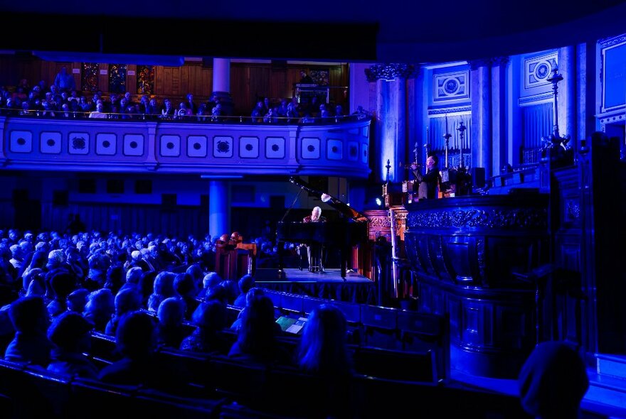 Concert on stage in a blue-lit auditorium with mezzanine and stalls seating.