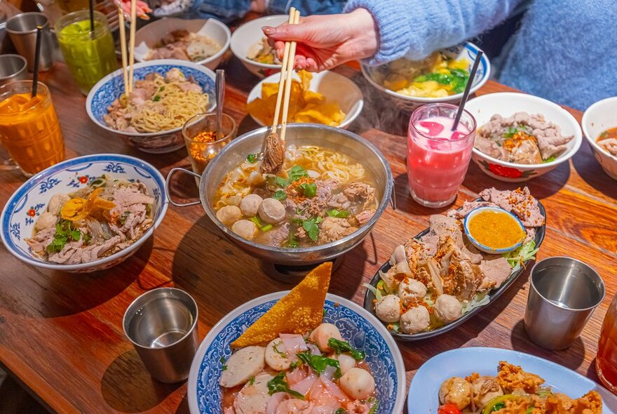 A table full of Thai food and a person is reaching into a bowl of noodles.