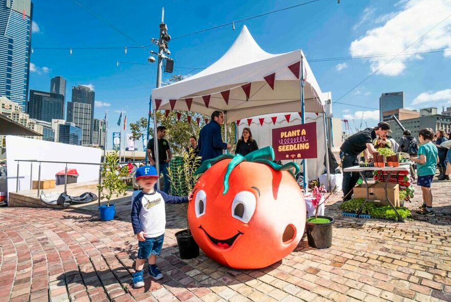 A child is standing next to a giant tomato mascot costume at a festival