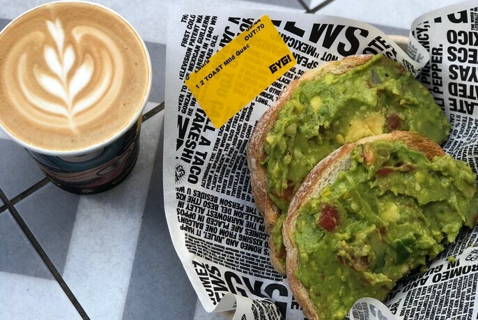 Avocado on toast in a newsprint wrapper and a cappuccino with stencil art. 