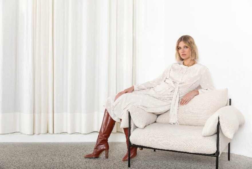 Model lounging on a seat, wearing a pale wool dress and long boots.