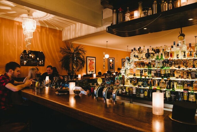 Bearded man sitting at a long wooden cocktail bar with candle lighting, taps and shelves filled with backlit bottles.
