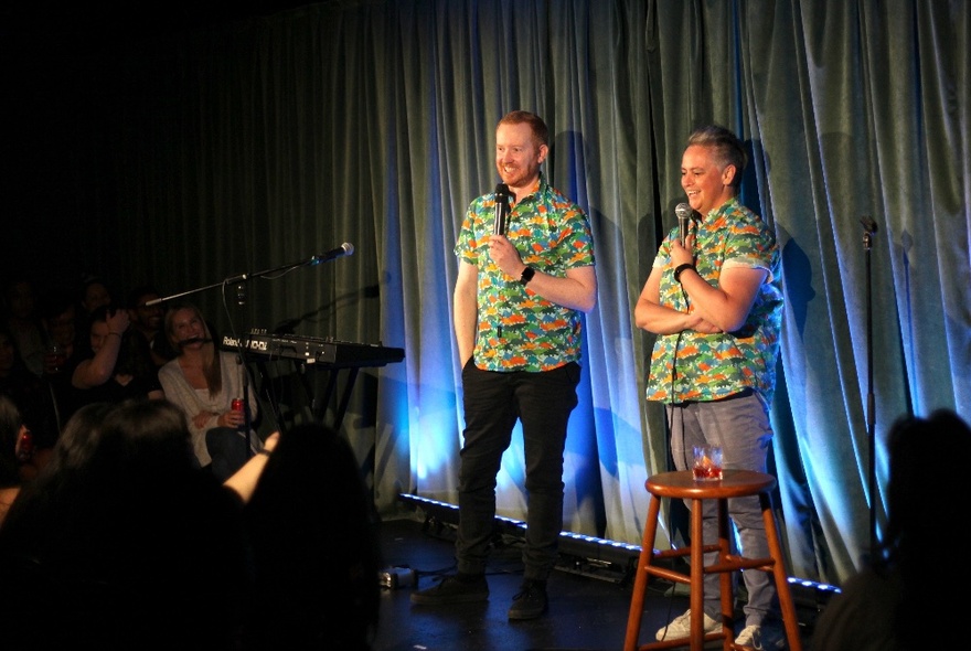 Two smiling comedians - Geraldine Hickey and Luke McGregor - on a small stage wearing matching shirts and holding microphones, with an audience watching.