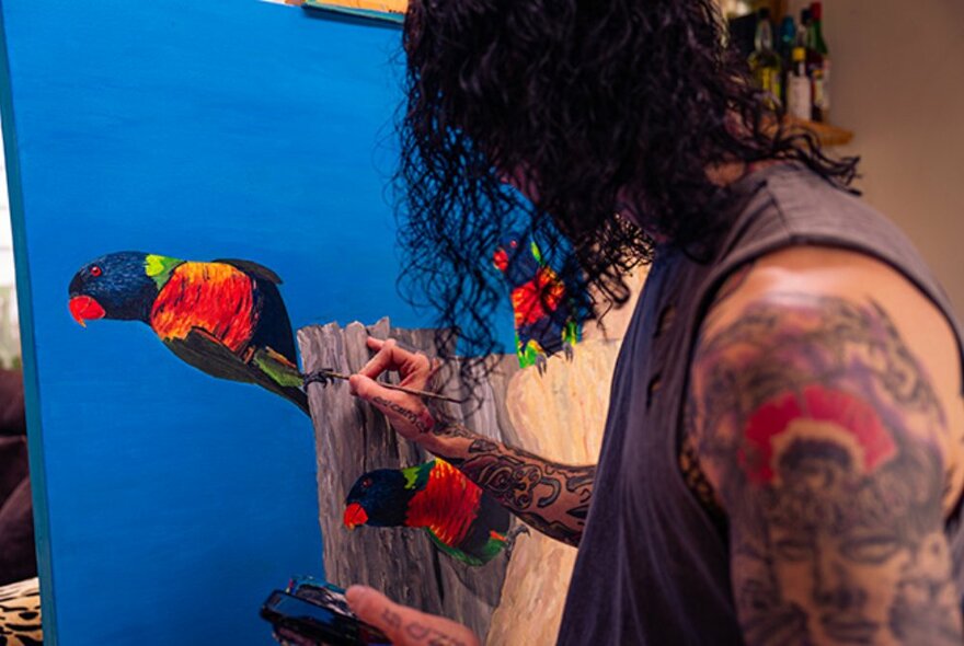 Person in a sleeveless vest, their arms decorated with tattoos, painting images of colourful birds against a bright blue sky onto a canvas.