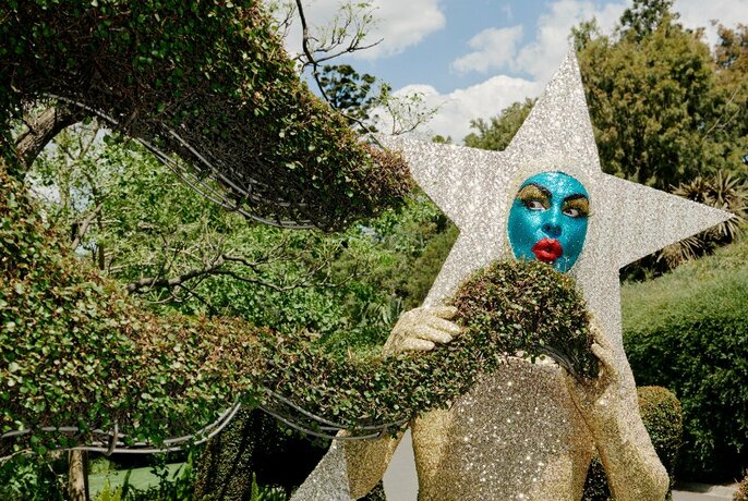 Person in a silver star costume with a painted blue face, posing among garden topiary.
