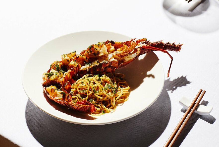 A half lobster and noodle dish presented in a white bowl with chopsticks to the side.