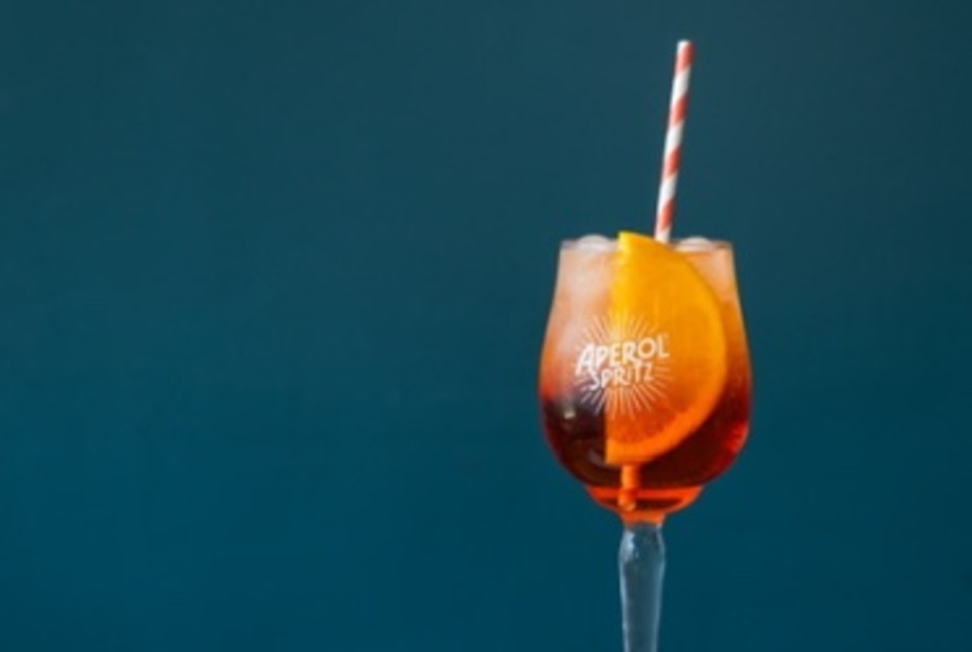 A single Aperol spritz with a striped straw against a deep blue background. 