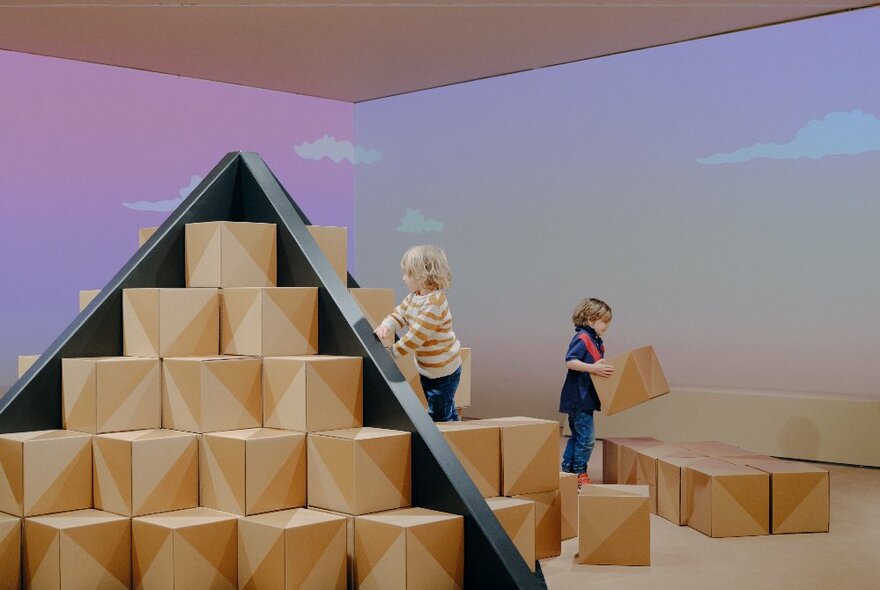 Two young children building a cardboard pyramid out of 3D cardboard shapes in a large gallery room.