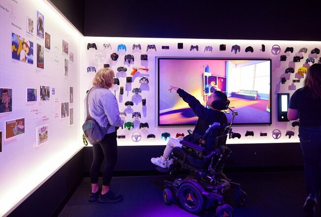 A person in a wheelchair pointing at a gallery display of video game controllers.