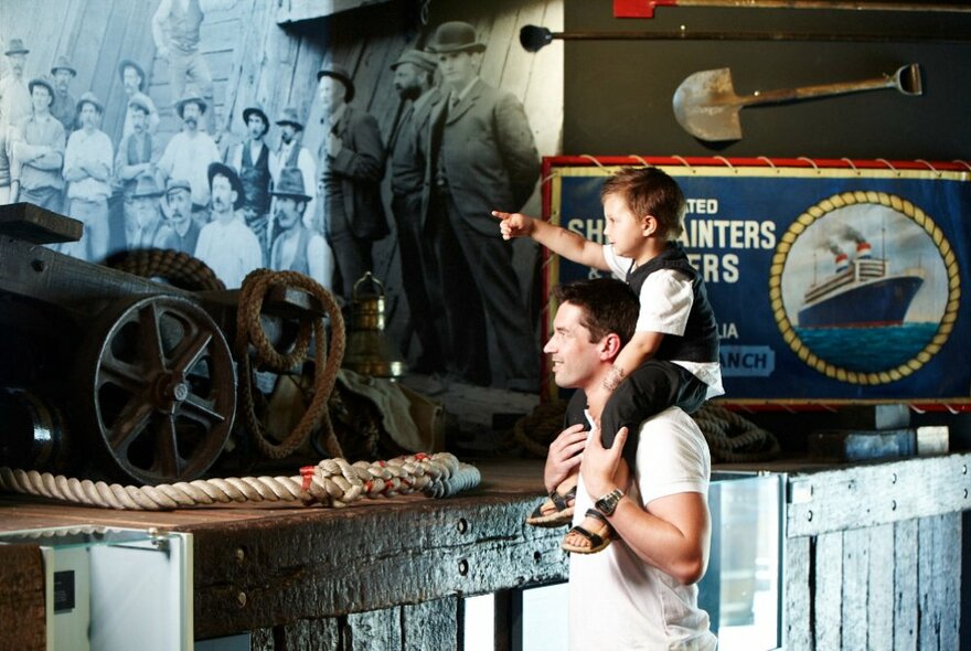 A young child sitting on the shoulders of an adult and pointing to a wall display in a maritime museum.