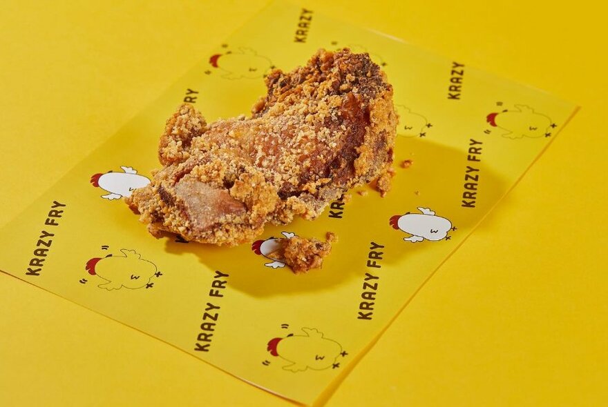 A crispy fried piece of chicken resting on a yellow piece of paper that is printed with the words 'KRAZY FRY' and line drawings of chickens. 