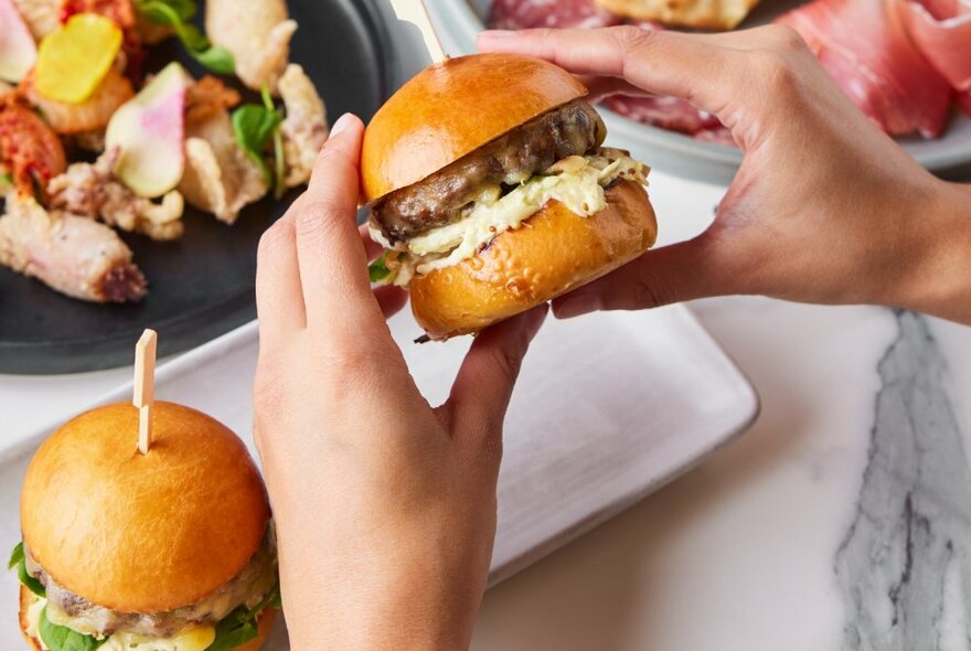 Hands holding a mini burger over a plate with another burger next to assorted dishes of food.
