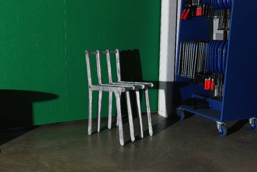 A chair crafted from solid aluminium rods, positioned against a green wall.