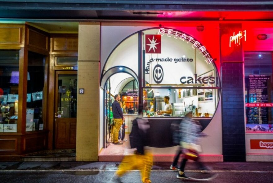 Pedestrians walking past a lit up gelato shop at night, the window is circular and there is a neon sign 