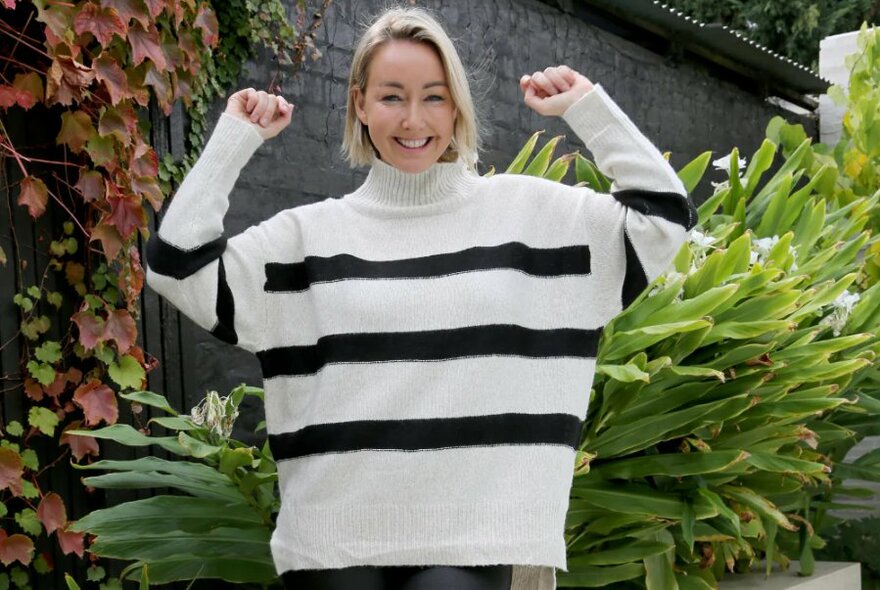 Smiling woman with a blond bob haircut wearing a white polo-necked jumper with black horizontal stripes across it, standing in an outdoor garden. 