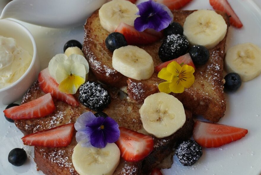 A serve of French toast on a white plate, garnished with slices of banana, strawberries, blueberries and icing sugar.